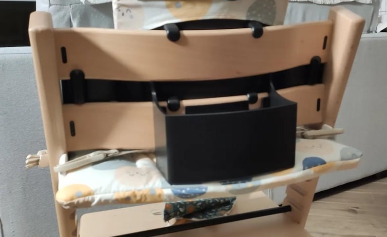 Puede incluir: Una trona de madera clara con una caja de almacenamiento negra. La silla tiene un coj&iacute;n estampado y est&aacute; dise&ntilde;ada para beb&eacute;s y ni&ntilde;os peque&ntilde;os. La silla es de madera.