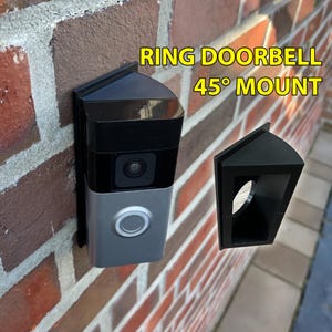 May include: A black 45-degree mount for a Ring Doorbell is displayed next to a mounted doorbell on a brick wall. The doorbell is silver and black. The text "RING DOORBELL 45° MOUNT" is in yellow.