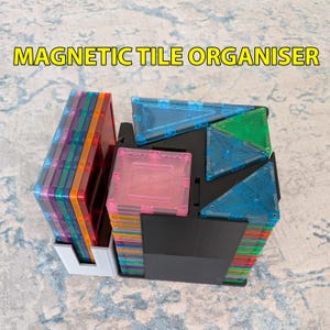 May include: A black and grey plastic magnetic tile organiser holding stacks of colourful translucent square and triangular magnetic tiles. The text MAGNETIC TILE ORGANISER is printed in yellow letters at the top of the image.