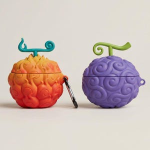 May include: Two silicone earbud cases shaped like stylised fruits with swirling patterns. The left case is a gradient of orange and red with a teal stem, and the right case is purple with a light green stem. A silver metal carabiner is attached to the left case.