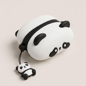May include: A white and black silicone protective case for wireless earbuds, shaped like a panda with black ears and eye patches. A small, matching panda charm hangs from a black cord attached to the side of the case, featuring pink cheeks.