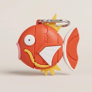 Puede incluir: Un llavero de silicona con forma de pez naranja estilizado con un ojo blanco, labios rosas, aletas amarillas y una cola blanca. El pez cuenta con un mosquetón de metal plateado en la parte superior para sujetarlo fácilmente a bolsos o llaves.