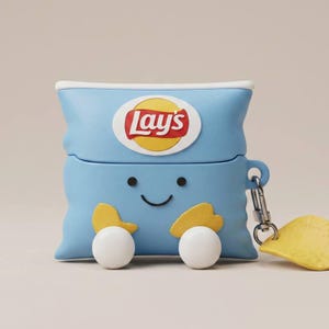May include: A light blue silicone protective case for wireless earbuds shaped like a Lay's potato chip bag. The case features a white oval logo with the red Lay's text, a smiling face, two yellow chip accents, two white spheres, and a metal carabiner.