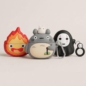 Puede incluir: Tres fundas de silicona para auriculares con diseños de personajes. La funda izquierda tiene forma de llama roja y amarilla. La central es una criatura gris con una pequeña figura blanca encima. La derecha es una figura negra con máscara blanca.