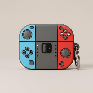May include: A protective silicone case for wireless earbuds designed to resemble a game console controller. The case features a central dark grey section with a light blue left side and a red right side, complete with directional buttons, joysticks, and a metal ring.