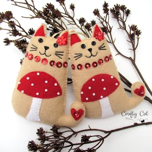 May include: Two handmade felt cat ornaments. The beige cats have red mushroom-shaped bodies, ear tips, and heart-shaped feet. They feature black eyes, sequin collars, and are decorated with red sequins and white dots. Small red flowers adorn the cats. The ornaments are for decoration.