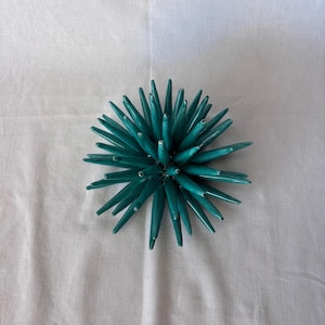 May include: A teal decorative object with a spiky, starburst design. The object has numerous pointed, elongated elements radiating outwards from a central point. It is set against a white background.