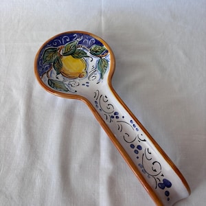 Puede incluir: Un reposacucharas de cerámica con base blanca, con un diseño pintado a mano de un limón amarillo con hojas verdes y detalles azules. El borde está delineado en un color marrón cálido. Utensilios de cocina decorativos.