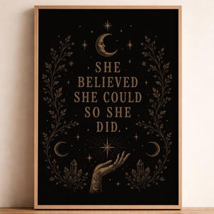 May include: A framed rectangular wall art print featuring a black background with gold-toned illustrations of a crescent moon, stars, crystals, and leafy vines. The text reads, "SHE BELIEVED SHE COULD SO SHE DID." A hand is depicted reaching toward a star.