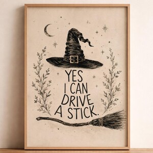 May include: A framed wall art print on a light beige background featuring a black ink illustration of a pointed witch hat, a broomstick, and floral sprigs. The text reads, YES I CAN DRIVE A STICK, surrounded by small stars and a crescent moon.