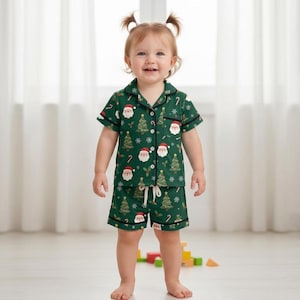 May include: A dark green short-sleeve pajama set featuring a repeating pattern of Santa Claus faces, decorated Christmas trees, snowflakes, and candy canes. The button-down shirt has a collar and a chest pocket, while the shorts have a drawstring waist.