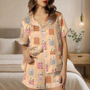 May include: A peach-colored pajama set with a floral and book print. The short-sleeved top and shorts feature a repeating pattern of floral arrangements in vases and books. White piping trims the edges of the set.