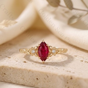 May include: A gold ring featuring a central marquise-cut red gemstone flanked by two small round white stones. The band is detailed with engraved leaf and floral motifs on both sides of the center setting, creating a botanical-inspired design.