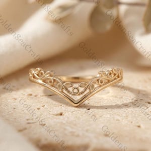 Può includere: Un anello color oro con un design a forma di V. L'anello presenta un intricato motivo a filigrana con dettagli floreali e a forma di foglia. L'anello è posto su uno sfondo neutro, evidenziando la sua delicata maestria. Il testo "GoldenSeaCrafts" è visibile.