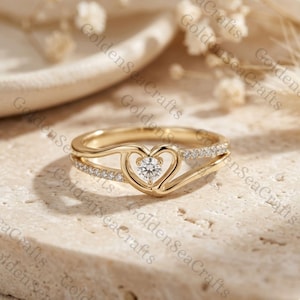 May include: A gold-colored ring with a heart-shaped design. The ring features a central, clear gemstone within the heart and a band with small, clear stones. The ring is set against a neutral background.