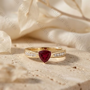 Può includere: Un anello in oro con una pietra preziosa rosso intenso taglio trillion al centro, affiancata da una fila di piccole pietre bianche rotonde incastonate su ciascun lato della fascia in oro lucido. L'anello è posizionato orizzontalmente.