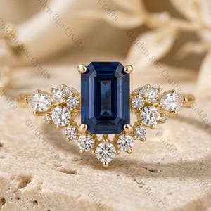 May include: A gold ring featuring a central emerald-cut dark blue gemstone flanked by a cluster of round and marquise-cut clear stones arranged in a curved, fan-like setting. The band is thin and metallic, showcasing a polished gold finish.