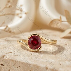 Pode incluir: Um anel de ouro com uma pedra preciosa redonda de cor vermelho profundo engastada numa banda de estilo bypass. O metal dourado polido curva-se em torno da pedra central, criando um design suave e contínuo que destaca a cor do rubi facetado.
