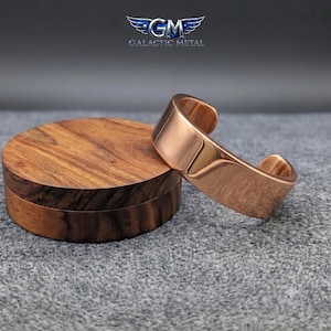 May include: A polished copper-colored cuff bracelet with a smooth, reflective surface and an open-ended design. The bracelet is positioned next to a circular wooden block with a visible grain pattern. The logo GM Galactic Metal is visible.