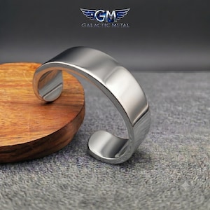 May include: A polished silver-toned metal cuff bracelet with a smooth, reflective surface and an open-ended design. The bracelet features a wide, curved band that maintains a consistent width throughout its circular shape, showcasing a clean finish.