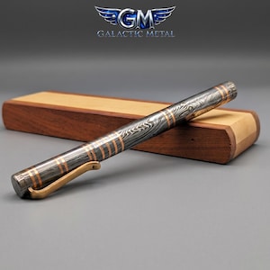 May include: A cylindrical pen featuring a dark gray and black wavy patterned metal body with thin copper-colored bands and a gold-toned metal clip. The pen rests in front of a rectangular wooden case with a dark brown base and a light tan top.