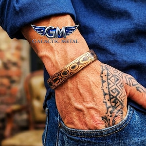 May include: A brown and tan metal cuff bracelet with a geometric pattern. The bracelet is worn on a wrist, with the Galactic Metal logo in the upper left corner. The person is wearing a blue denim shirt.
