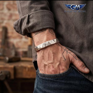 Puede incluir: Un brazalete de metal en tono plateado con una textura martillada y facetada. El brazalete presenta un diseño de banda ancha y sólida con un acabado pulido que refleja la luz sobre su superficie angular y geométrica, usado en la muñeca.