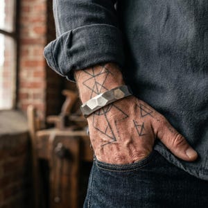 May include: A wide, metallic cuff bracelet with a faceted, geometric surface texture. The bracelet has a matte, brushed finish and a solid, rigid construction that encircles the wrist, showcasing a series of angular, multi-sided planes.