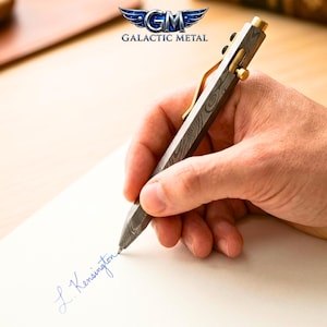 May include: A bolt-action pen with a Damascus steel barrel featuring a swirling gray and black pattern. The pen includes a gold-colored metal pocket clip and a matching gold-colored bolt mechanism. The top of the pen is capped with a gold-colored button.