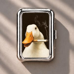 May include: A rectangular silver metal cigarette case featuring a high-resolution print of a white duck with an orange beak holding a lit cigarette in its mouth, with a thin trail of grey smoke rising against a dark brown background.
