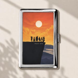 May include: A rectangular silver metal case featuring a printed graphic of seven silhouettes standing on a cliff edge against an orange sunset sky with a bright sun. The text MIGHTY NEIN is printed in white below the figures, with mountains visible below.