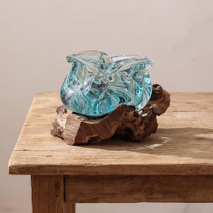 Könnte beinhalten: Eine dekorative, hellblaue Glasschale mit einem strukturierten, wirbelnden Design. Die Schale steht auf einem Stück natürlichem, dunkelbraunem Holz, was einen Kontrast in Texturen und Farben erzeugt. Die Form der Schale ist organisch und fließend.