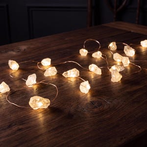 Op de afbeelding: Een LED-lichtsnoer met ruwe, doorschijnende, gebroken witte, kristalachtige stenen bevestigd aan een dun koperdraad. De lampjes verspreiden een warme, zachte gloed die de onregelmatige, grillige vormen van de stenen langs het snoer verlicht.
