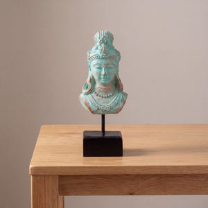 Op de afbeelding: Een turquoise buste van een godheid met gesloten ogen, versierd met een gedetailleerde hoofdtooi en sieraden. De sculptuur is gemonteerd op een zwarte standaard, wat een opvallend contrast creëert. De buste staat op een licht houten tafel.
