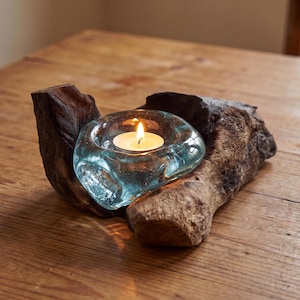 May include: A teal glass candle holder with a lit white candle, nestled in a piece of weathered driftwood. The wood has a rough, textured surface and a dark brown color. The candle flame casts a warm glow.