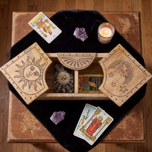 May include: A rectangular wooden box with two hinged lids featuring engraved sun and moon designs, containing two decks of tarot cards. The set includes three tarot cards placed outside the box, two amethyst crystal clusters, and a lit candle.