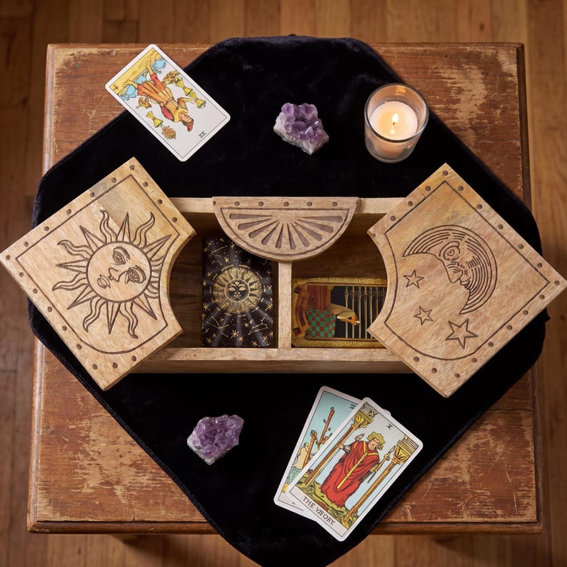 May include: A rectangular wooden box with two hinged lids featuring engraved sun and moon designs, containing two decks of tarot cards. The set includes three tarot cards placed outside the box, two amethyst crystal clusters, and a lit candle.