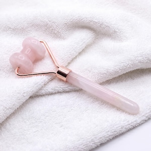 Pode incluir: Um rolo facial de cabeça dupla com dois rolos de quartzo rosa claro fixados numa estrutura de metal em ouro rosa, com um cabo cilíndrico liso de quartzo rosa claro. Esta ferramenta foi concebida para massagens faciais e cuidados da pele.