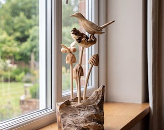 May include: A light brown wooden sculpture featuring a carved bird perched on a textured branch above several carved mushroom shapes and a small bee figure, all mounted on a solid, rough-hewn wooden base with a natural, light wood grain finish.