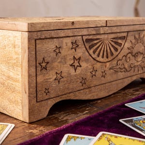 May include: A rectangular wooden box featuring a natural light brown grain and a hinged lid. The front panel is decorated with laser-engraved designs, including multiple five-pointed stars, a semi-circular sunburst, and a sun partially obscured by clouds.