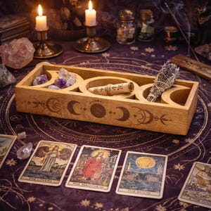 May include: A rectangular wooden tray featuring carved moon phase symbols on the front. The tray contains several small polished crystals, a rolled parchment scroll tied with twine, and a bundle of dried lavender and sage herbs.