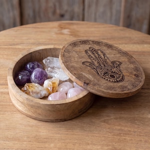 Peut inclure: Une boîte ronde en bois avec un couvercle amovible orné d'un symbole de main Hamsa gravé au laser. Le récipient ouvert contient une collection de pierres précieuses polies et brutes, dont de l'améthyste violette, du quartz clair, de la citrine jaune et du quartz rose.