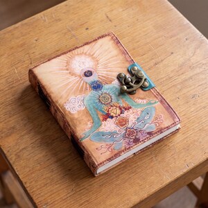 May include: A rectangular journal with a tan leather cover featuring a printed illustration of a figure in a seated meditation pose with visible chakra symbols. The journal is secured with a bronze-colored metal latch on a teal leather strap.