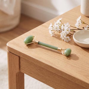 Pode incluir: Um rolo facial de extremidade dupla com duas pedras de jade verde-claro lisas, fixadas a um cabo central por ferragens de metal dourado. O rolo tem uma pedra oval grande numa extremidade e uma pedra oval mais pequena na outra extremidade.