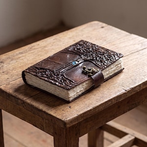 Könnte beinhalten: Ein dunkelbraunes Lederjournal mit geprägtem Baummotiv auf dem Einband, einer zentralen blauen, augenförmigen Einlage und einem messingfarbenen Metallverschluss. Das Journal hat dicke, strukturierte, cremeweiße Papierseiten an den Rändern.