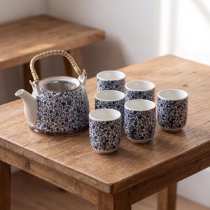 Może przedstawiać: Ceramiczny zestaw do herbaty składający się z okrągłego czajnika z plecionym bambusowym uchwytem i zaparzaczem ze stali nierdzewnej oraz pięciu pasujących filiżanek. Każdy element ma białą podstawę z gęstym, niebieskim wzorem roślinnym.