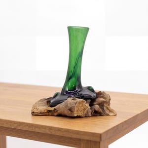 Op de afbeelding: Een groene glazen vaas met een uitlopende bovenkant en een donkere basis, gemonteerd op een stuk natuurlijk drijfhout. De vaas heeft een gladde, glanzende afwerking. Het staat op een lichtbruine houten tafel.
