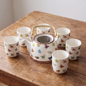 May include: A white ceramic tea set featuring a teapot with a woven bamboo handle and a stainless steel mesh infuser, accompanied by five matching cups. Each piece is decorated with a colourful pattern of various butterflies in shades of orange, blue, yellow, and green.