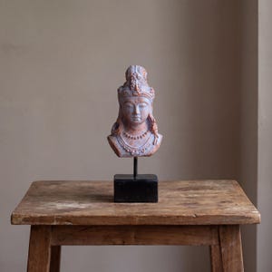 May include: A detailed bust of a serene figure, featuring an ornate headdress and jewellery, mounted on a black stand. The sculpture has a muted colour palette of blue, red, and white. It is displayed on a rustic wooden table.
