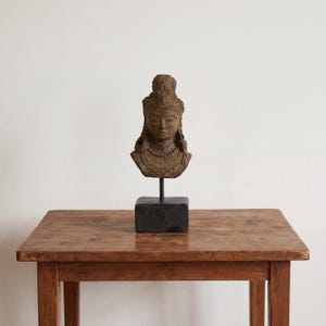 Op de afbeelding: Een gedetailleerde buste van een godheid, met ingewikkelde gravures en een serene uitdrukking. De sculptuur is gemonteerd op een zwarte rechthoekige basis met een zwarte metalen staaf. De buste heeft een warme, aardse tint en staat op een houten tafel.